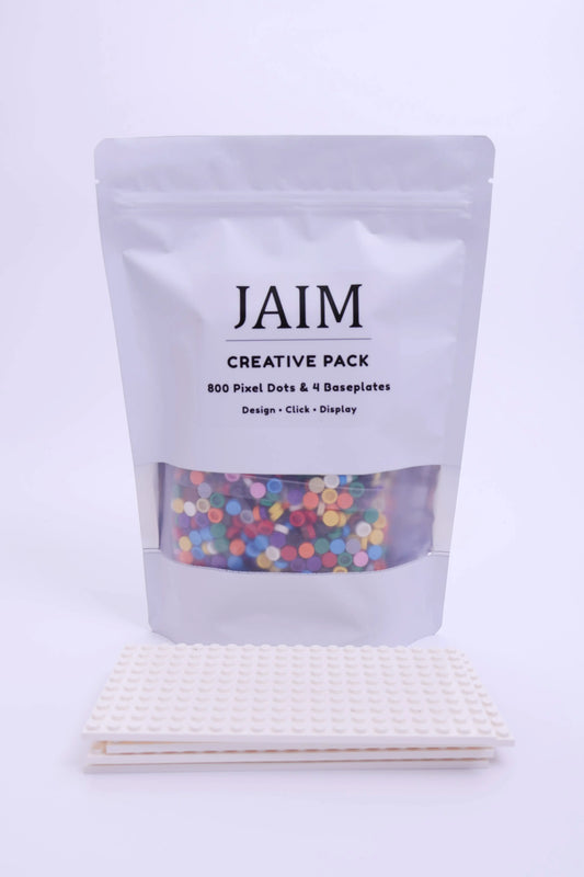A white stand-up pouch labeled 'JAIM CREATIVE PACK' with a clear window showing multicolored pixel dots, placed above two white pixel art baseplates on a clean white background.